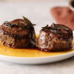 Two seared filet mignon steaks on a serving plate with crispy sage on top