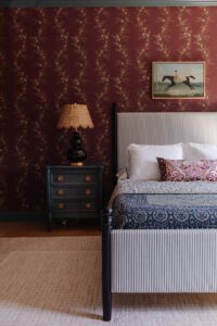 Vintage bedroom with dark floral wallpaper, striped upholstered bed, black bedside table with antique lamp, and horse riding artwork, showcasing traditional and eclectic interior decor.
