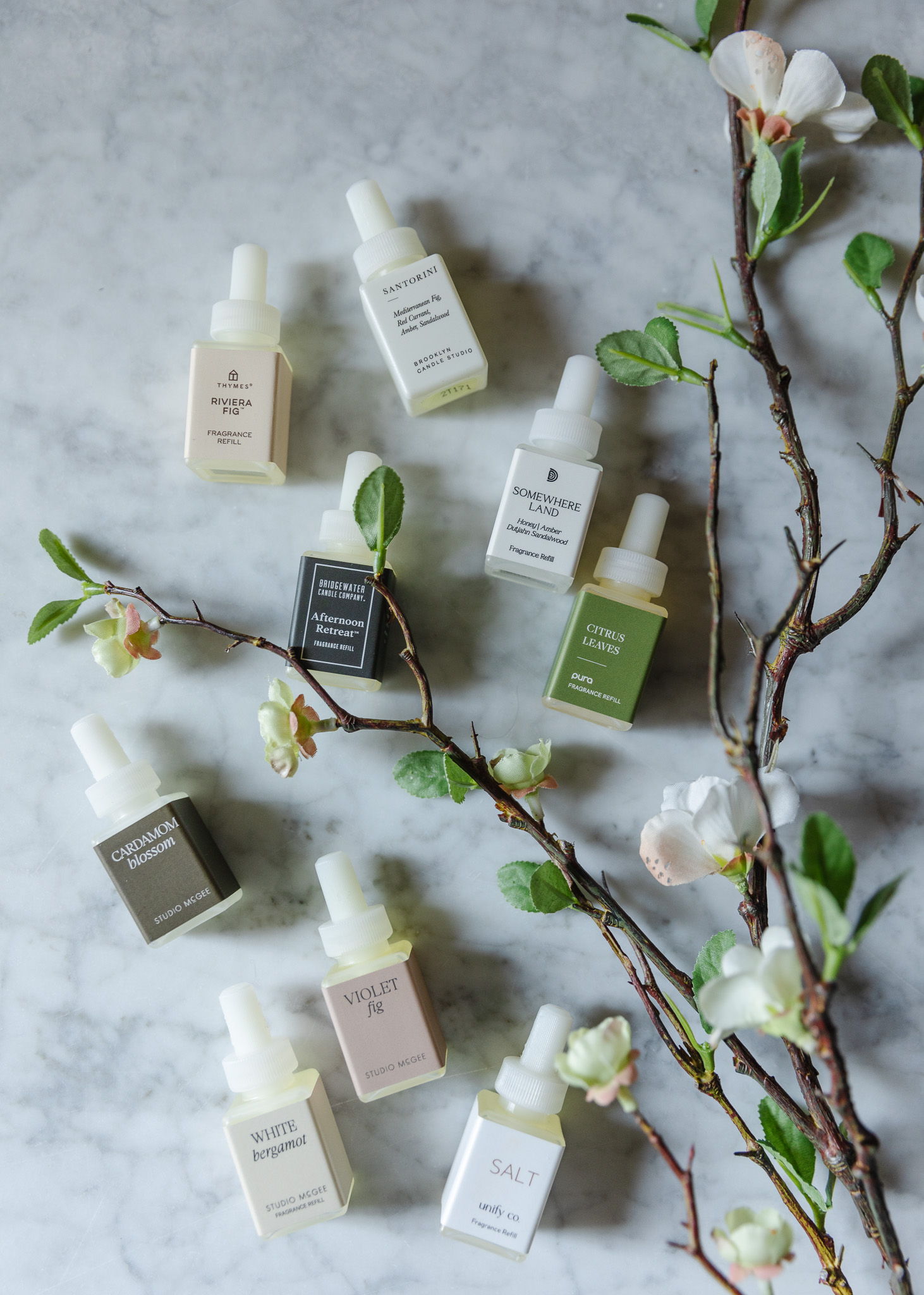 A flat-lay of nine Pura bottles on a marble countertop next to a branchy faux stem