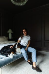 Julia in a white tee and jeans sitting on a bench at the foot of her bed with Cricket at her side