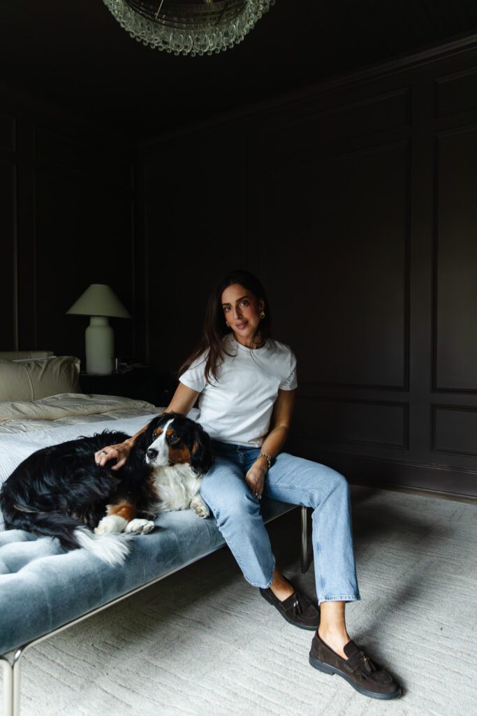 Julia in a white tee and jeans sitting on a bench at the foot of her bed with Cricket at her side