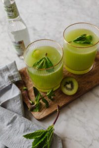 Two glasses of kiwi cucumber spritzer next to some cut kiwi and mint leaves