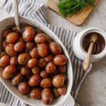 A white casserole dish with roasted red baby potatoes next to a honey jar and cutting board of fresh dill