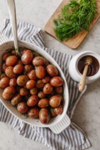 A white casserole dish with roasted red baby potatoes next to a honey jar and cutting board of fresh dill