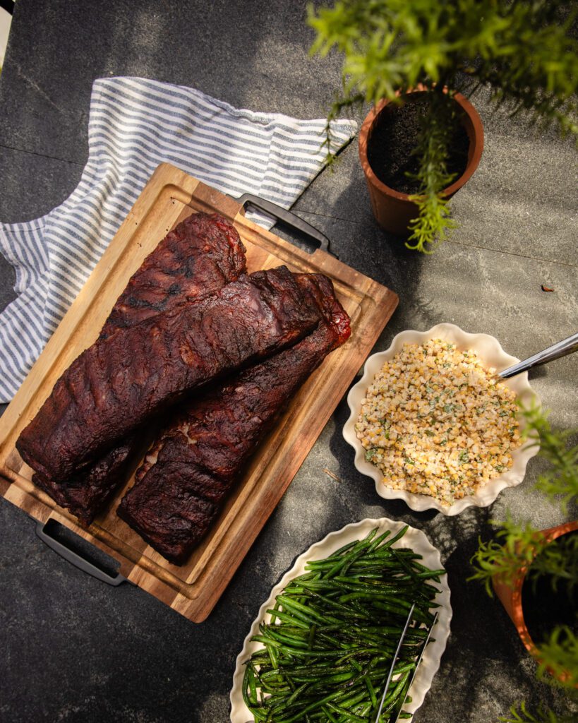 Two racks of smoked ribs with barbecue sauce on a wood cutting board next to green beans and corn salad