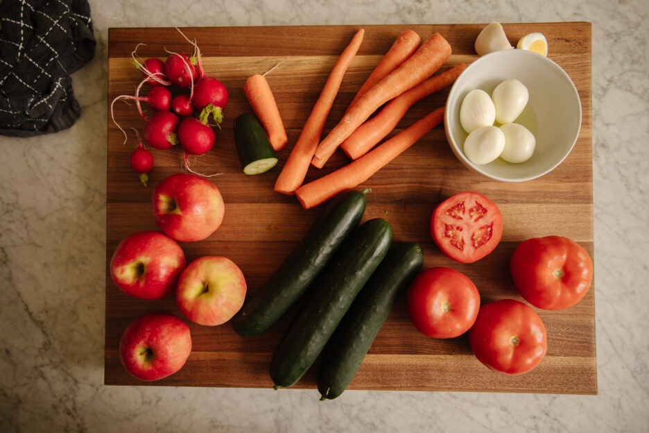 A cutting board with carrots, cucumbers, apples, tomatoes, hard-boiled eggs, and radishes