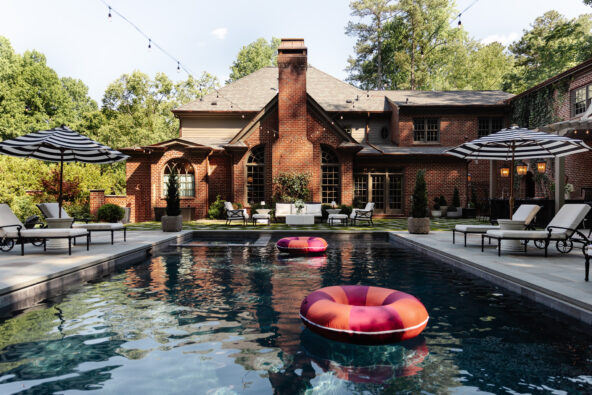 A spacious backyard featuring a rectangular swimming pool with two colorful pool floats, surrounded by lounge chairs and umbrellas, with a large brick house and lush greenery in the background.
