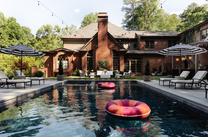 A spacious backyard featuring a rectangular swimming pool with two colorful pool floats, surrounded by lounge chairs and umbrellas, with a large brick house and lush greenery in the background.