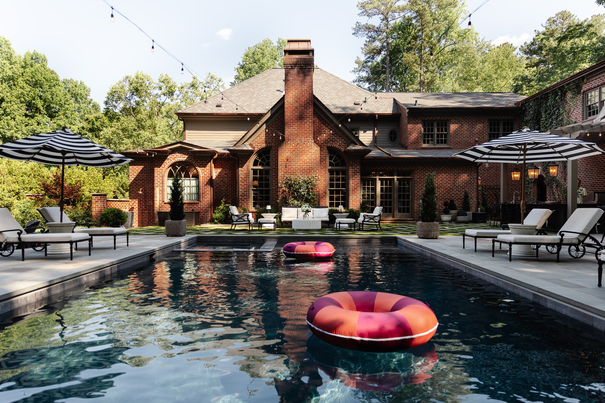 A spacious backyard featuring a rectangular swimming pool with two colorful pool floats, surrounded by lounge chairs and umbrellas, with a large brick house and lush greenery in the background.