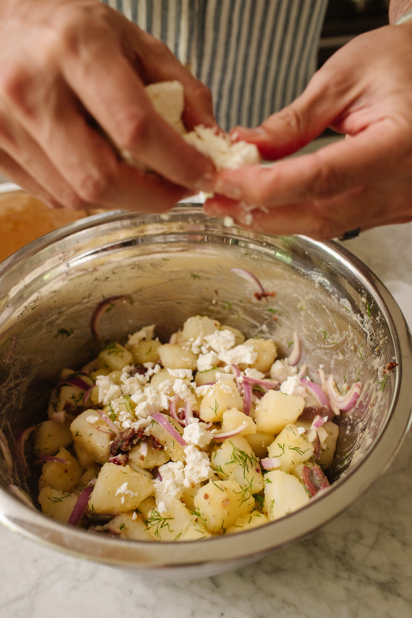 Hands crumbling feta into a bowl of cooked potatoes