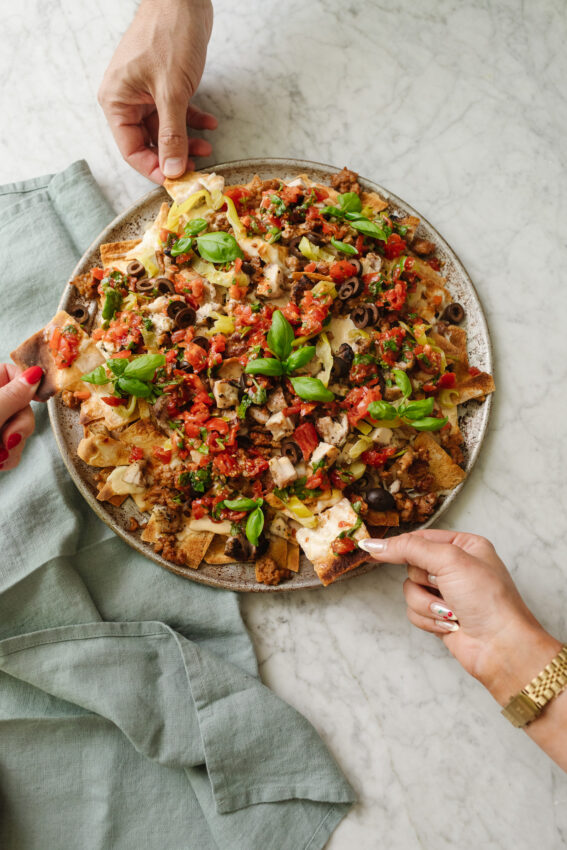 A platter of warm, loaded Italian nachos with three hands digging in to grab a chip