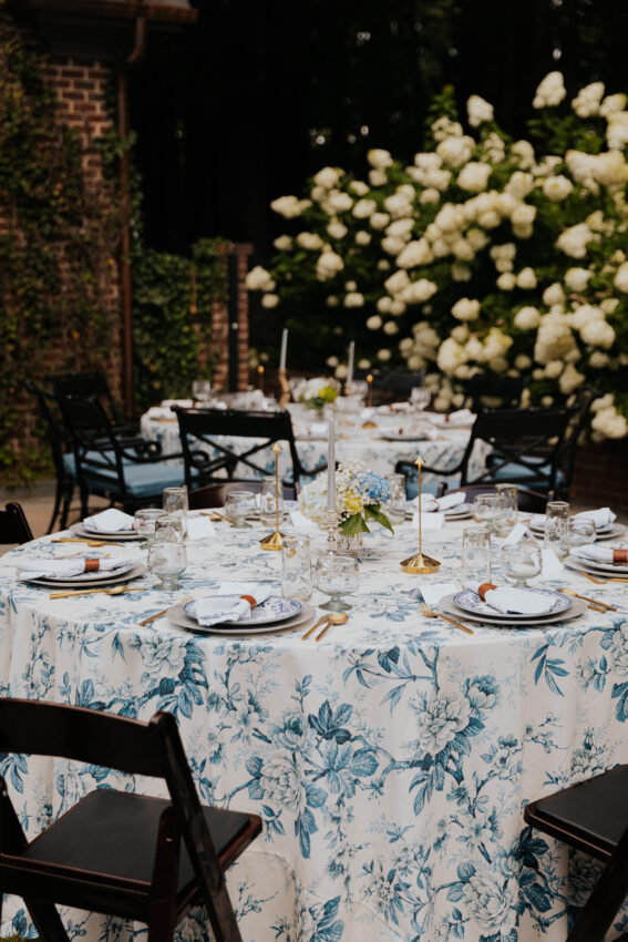 Outdoor round tables set for a dinner party with blue and white floral tablecloths, table lamps, and candles