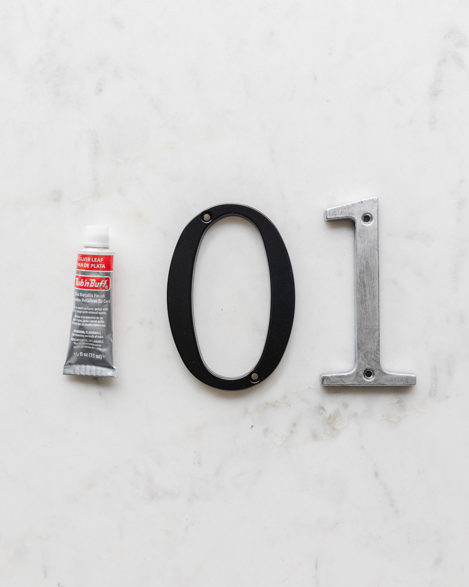 A tube of Silver Leaf Rub 'n Buff next to a plain black metal house number and a Rub 'n Buff painted house number