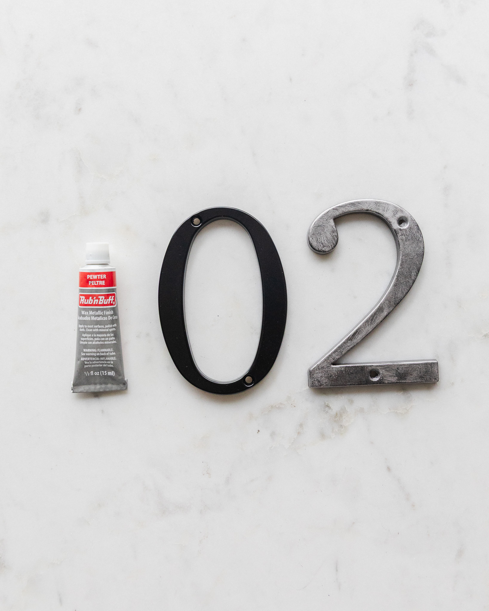 A tube of Pewter Rub 'n Buff next to a plain black metal house number and a Rub 'n Buff painted house number