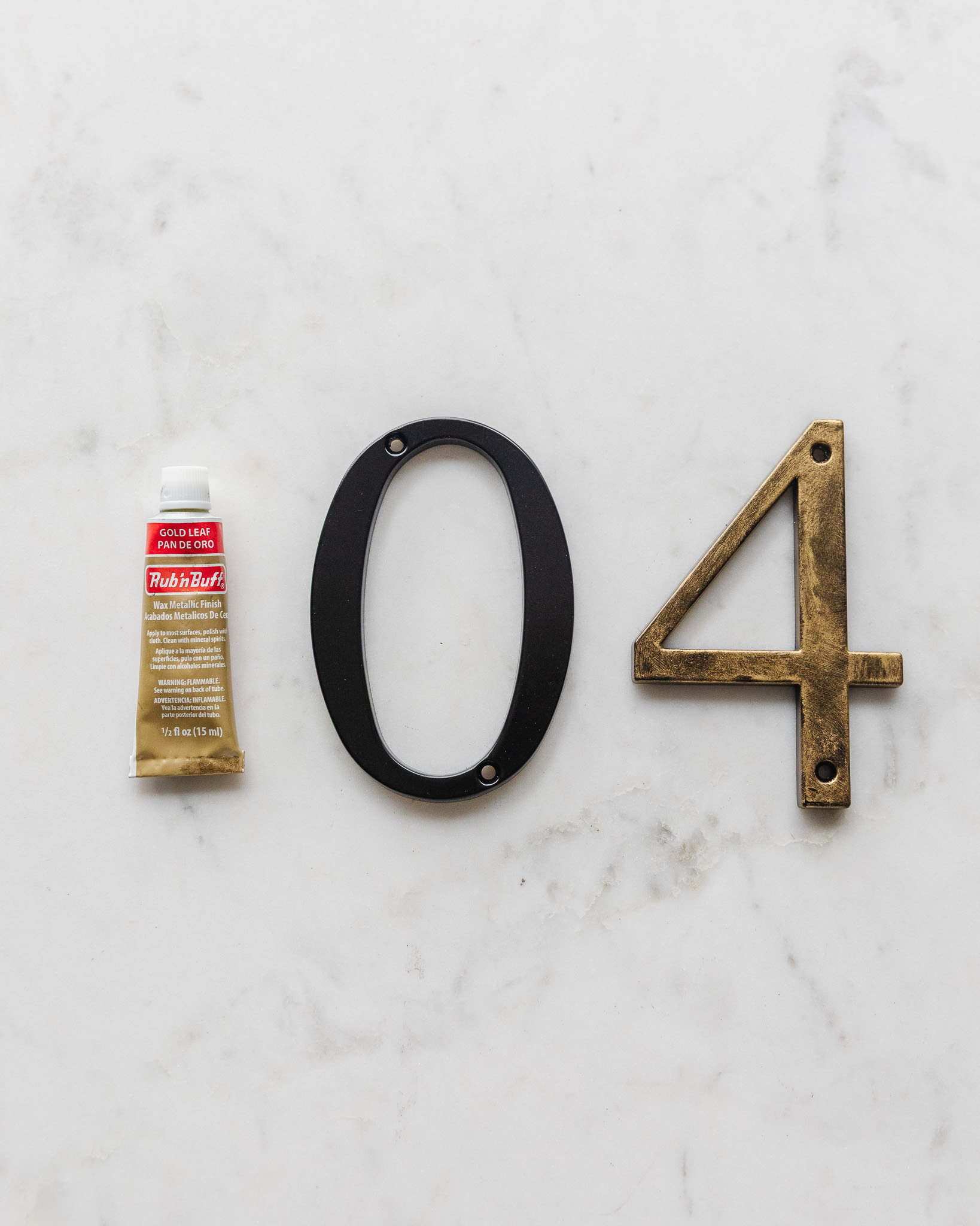 A tube of Gold Leaf Rub 'n Buff next to a plain black metal house number and a Rub 'n Buff painted house number