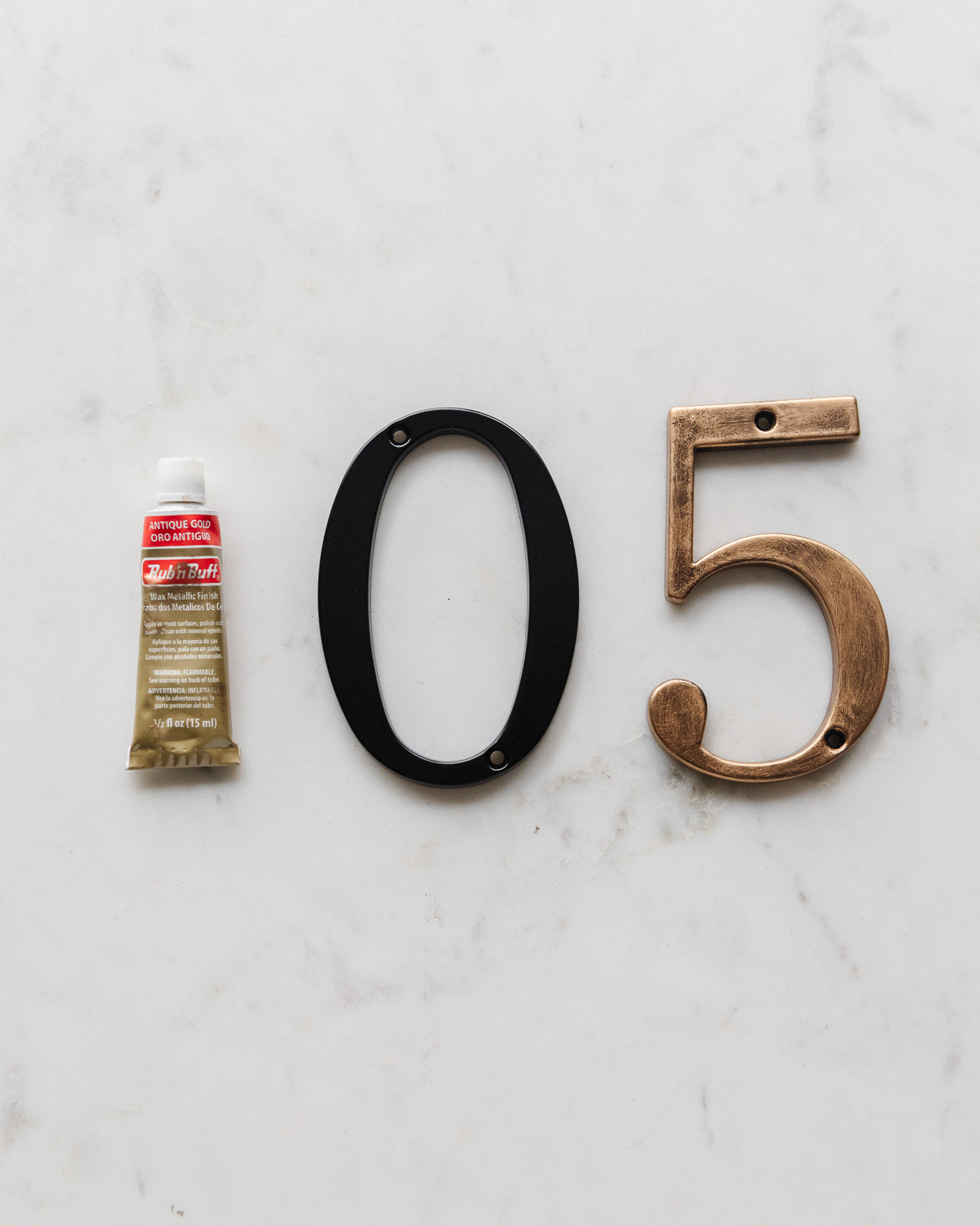 A tube of Antique Gold Rub 'n Buff next to a plain black metal house number and a Rub 'n Buff painted house number