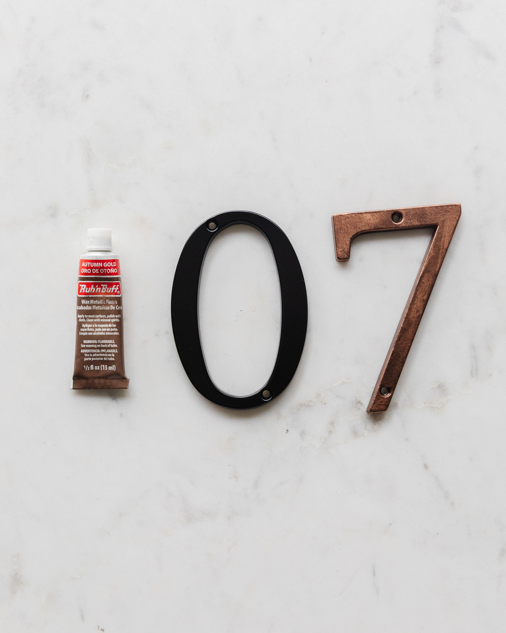 A tube of Autumn Gold Rub 'n Buff next to a plain black metal house number and a Rub 'n Buff painted house number