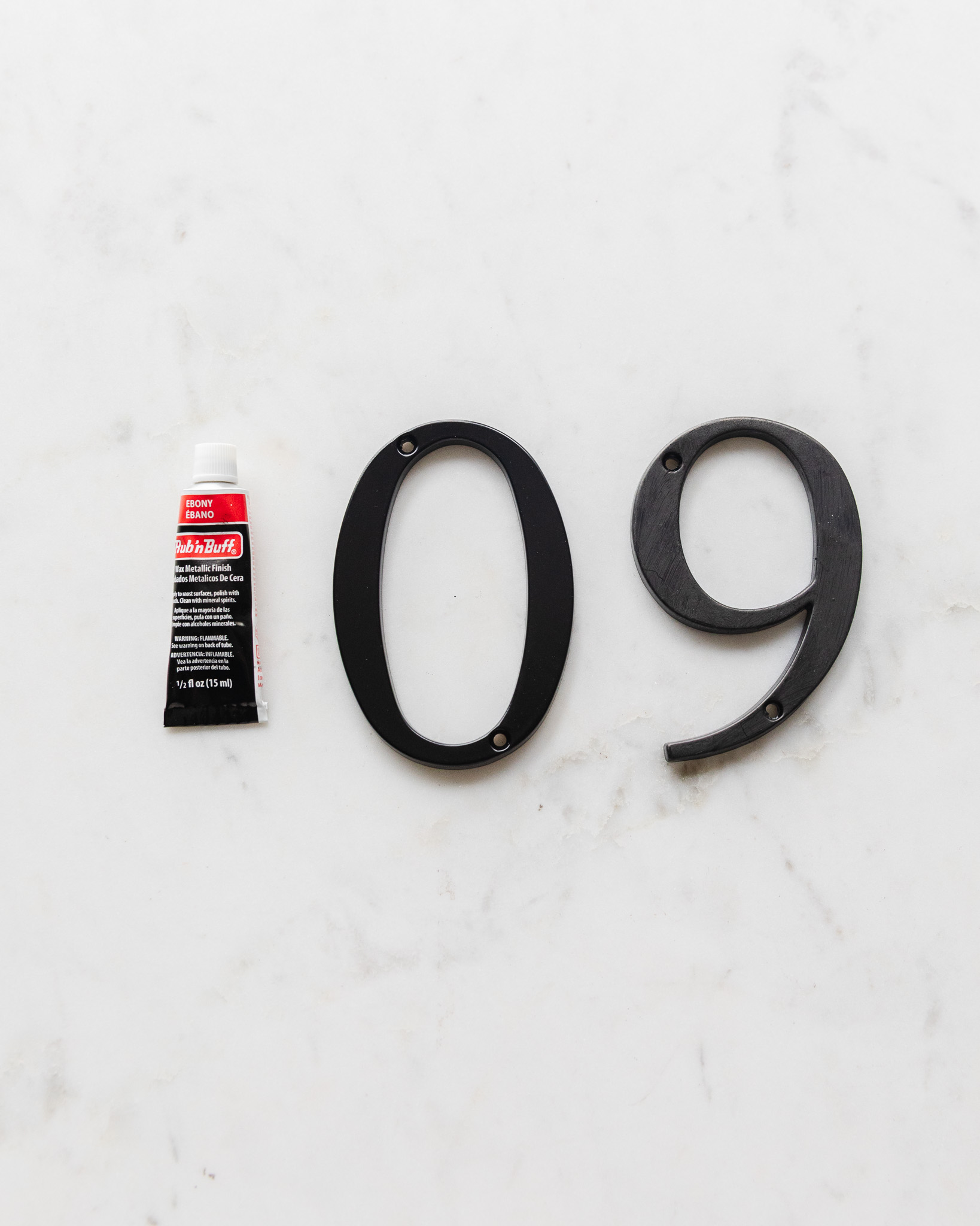 A tube of Ebony Rub 'n Buff next to a plain black metal house number and a Rub 'n Buff painted house number