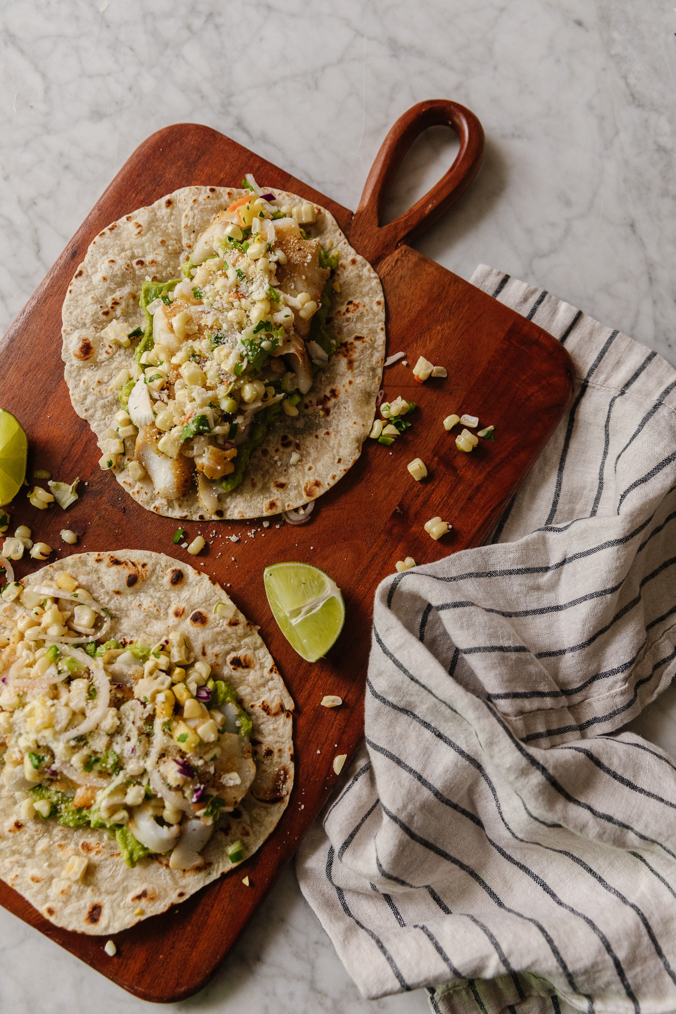 Cutting board with two fish tacos on it and lime wedges next to it