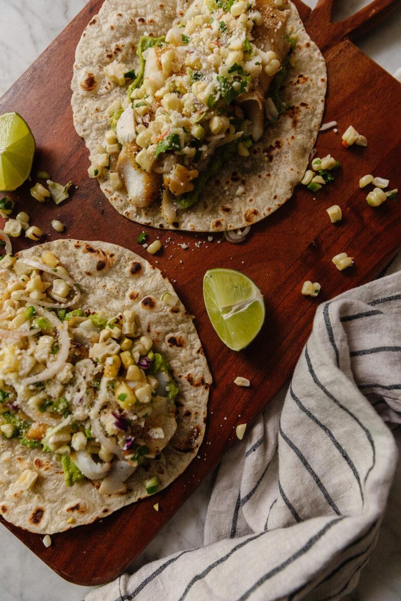 Cutting board with two fish tacos on it and lime wedges next to it