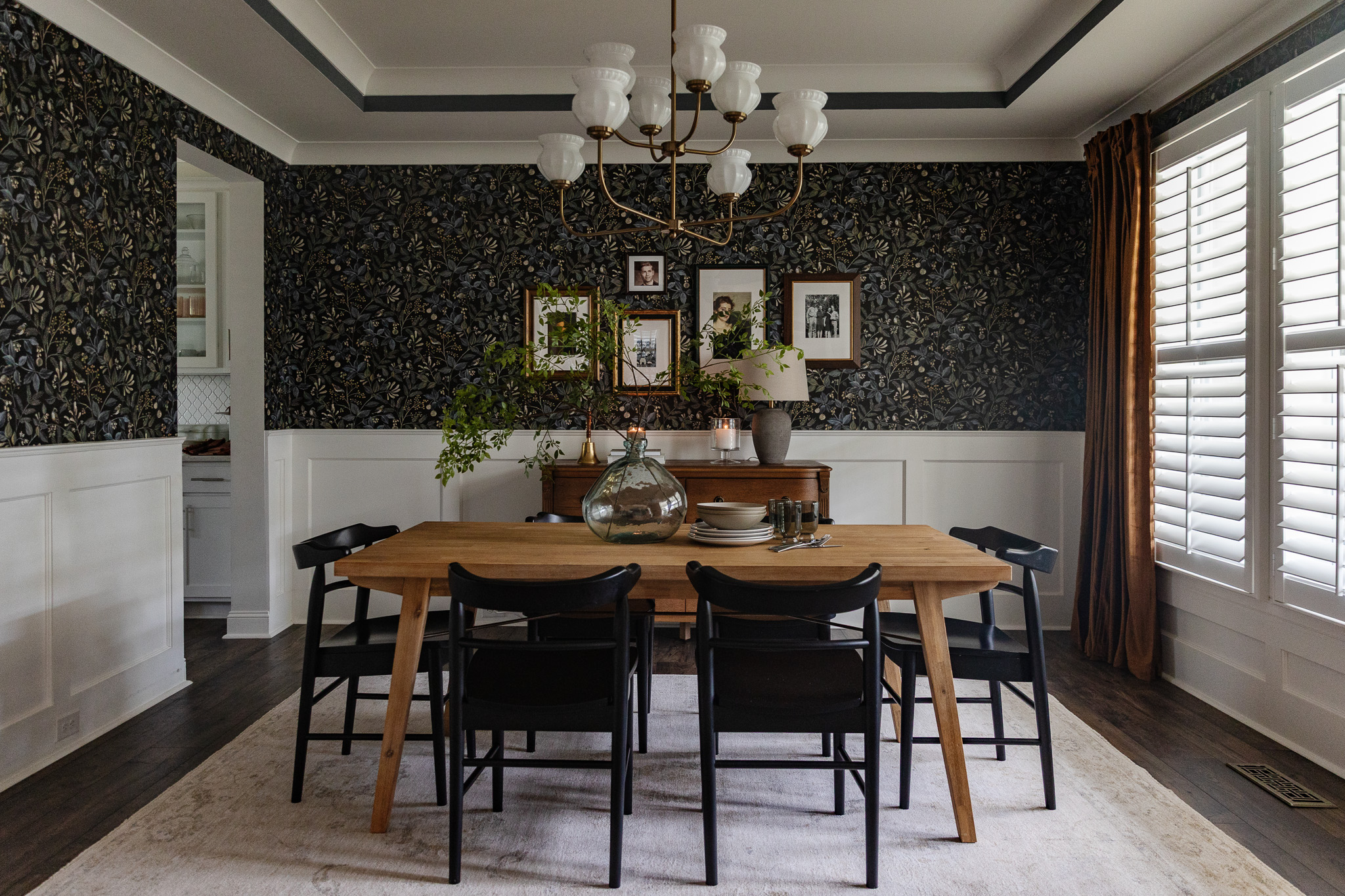 A blue dining room with dark floral wallpaper, black chairs and a vintage-modern chandelier