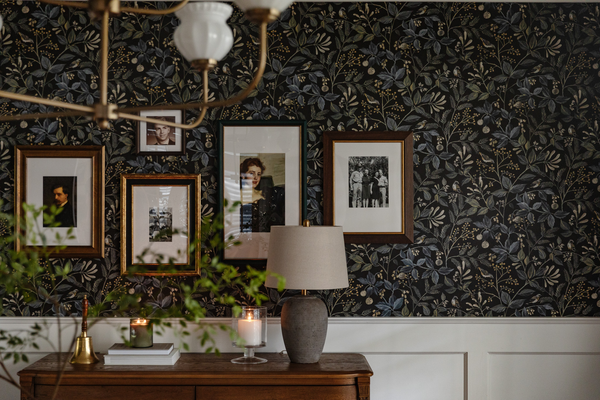 Close-up of the dark floral peel-and-stick wallpaper with a gallery wall of frames on top and a brown lamp on a sideboard