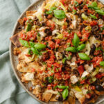 Overhead of a platter of Italian nachos featuring sausage, chicken, olives, pepproncini, mozzerella cheese, basil, and chopped tomatoes