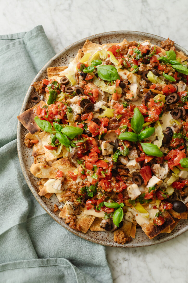 Overhead of a platter of Italian nachos featuring sausage, chicken, olives, pepproncini, mozzerella cheese, basil, and chopped tomatoes