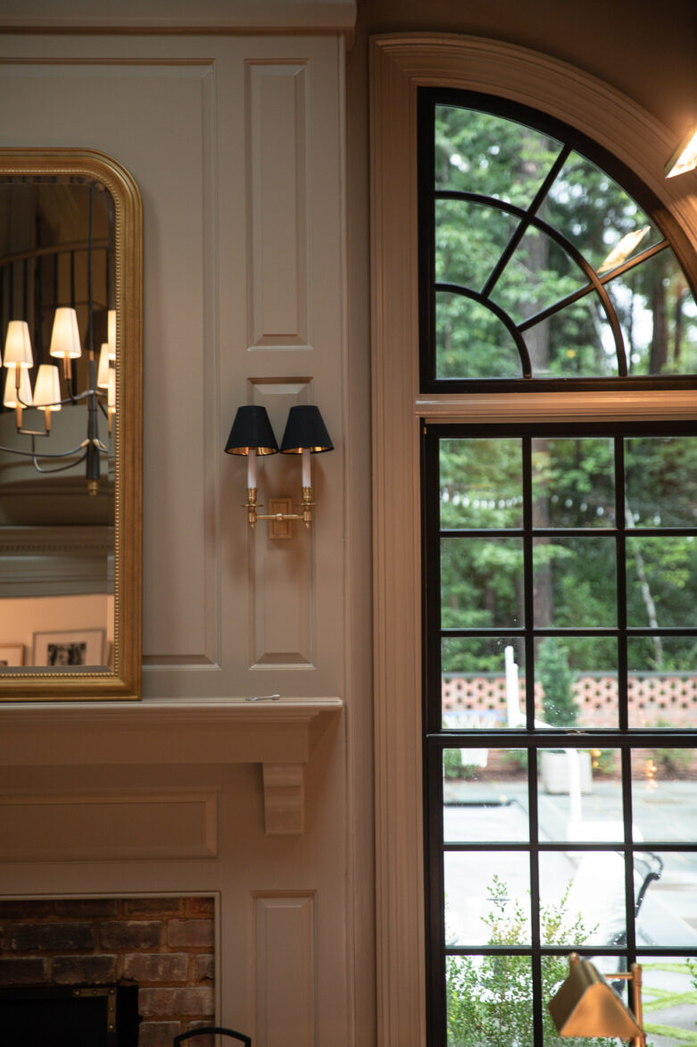 A gold double shade sconce with black shades beside the fireplace mantle and mirror