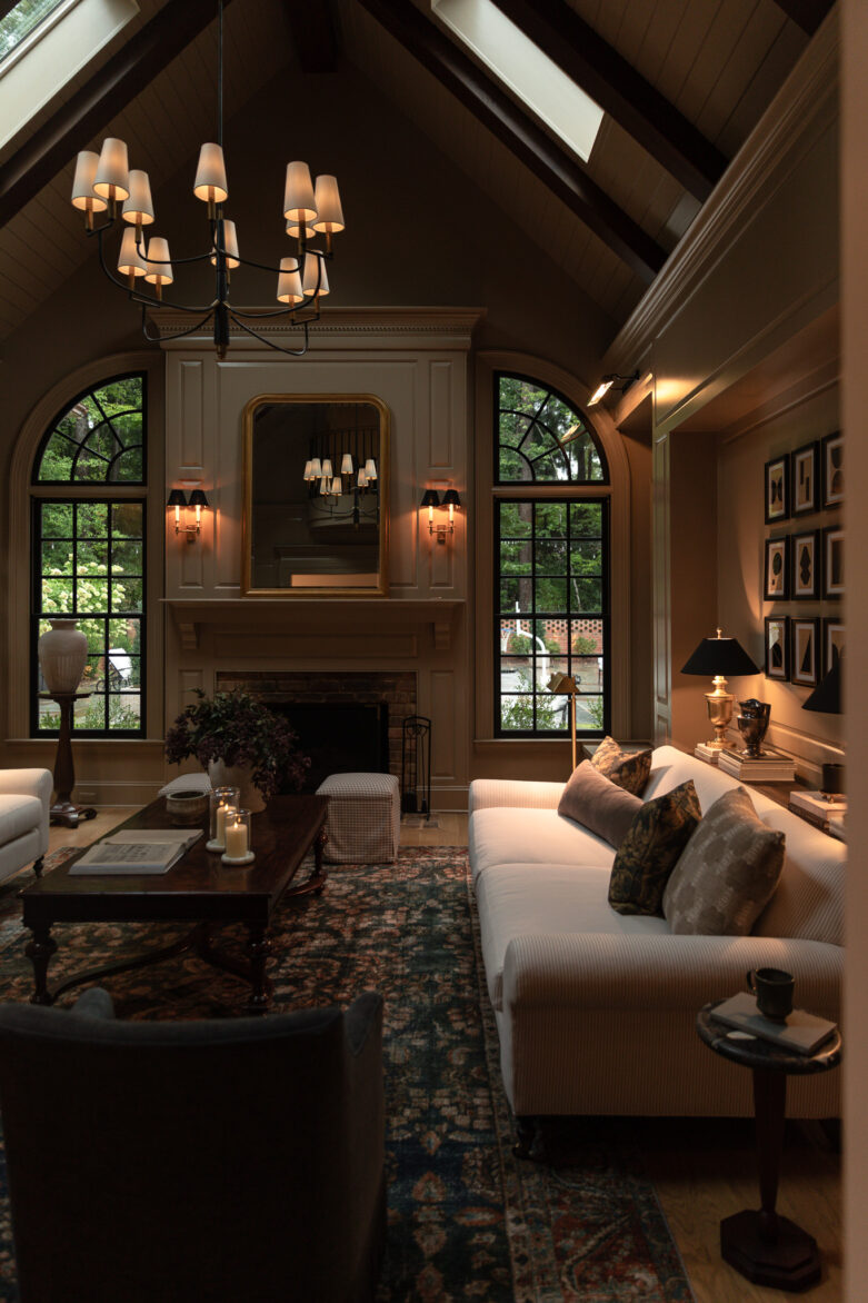A moody, inviting view of a living room with vaulted ceilings, dark brown color palette, and glowing lamps, sconces, and chandelier. Lamp 'o clock at its finest.