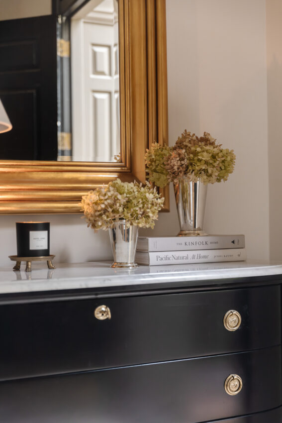 Elegant entryway featuring a black dresser with gold accents, topped with two glass vases filled with hydrangeas, and a large gold-framed mirror reflecting a doorway.