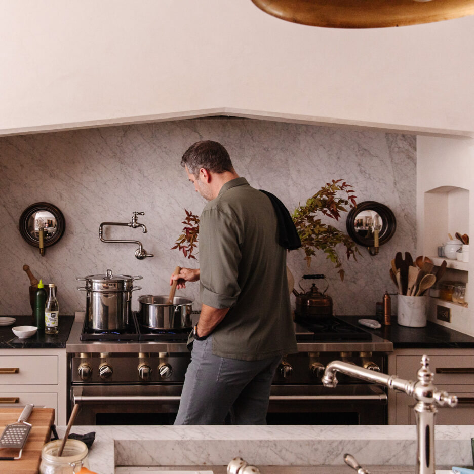 Modern kitchen with stainless steel appliances and a marble backsplash, featuring a man cooking on the stove, stylish cabinetry, and decorative wall accents for a warm, inviting home cooking space.