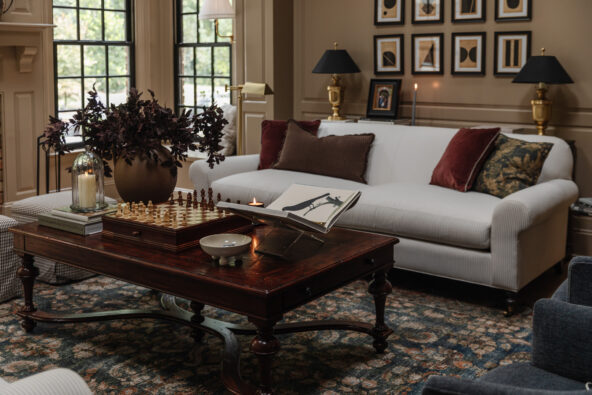 Elegant living room with a plush white sofa, decorative pillows, and a cozy patterned rug, featuring classic wall art, table lamps, and a central wooden coffee table adorned with books, a chess set, and floral accents.