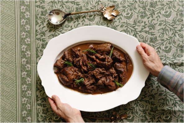 Slow-cooked lamb served on a white platter, garnished with rosemary sprigs, on a green patterned tablecloth, featuring a vintage silver spoon with a flower-shaped handle.