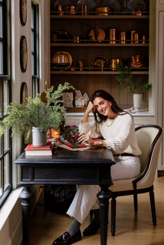 Cozy kitchen nook with holiday decor, featuring a woman reading a holiday card at a dark wood table near a window with greenery and festive accents, showcasing warm, inviting home design.