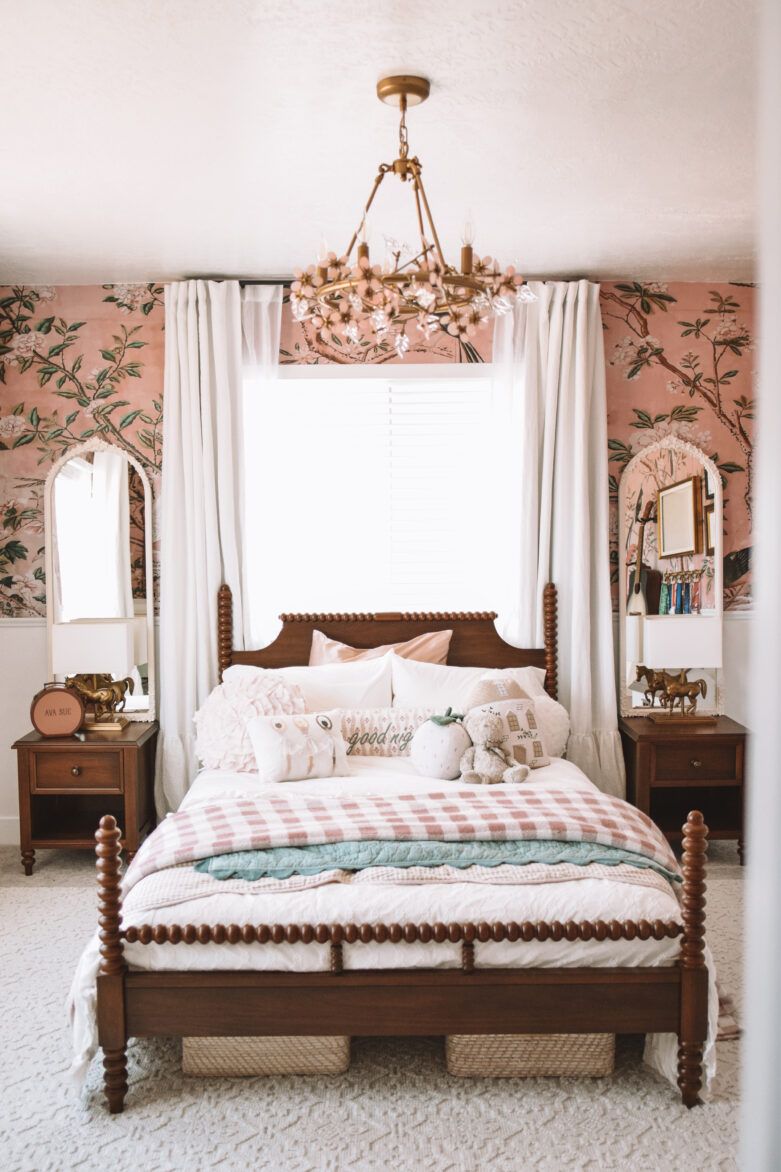Vintage wooden bed with tufted bedding and plush pillows in a dreamy, floral-themed bedroom with pink wallpaper.