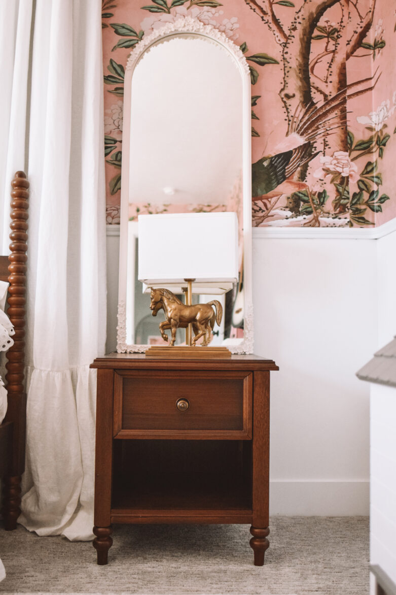 Vintage wooden nightstand with a mirror and a gold horse table lamp, set against pink floral wallpaper in a bedroom.