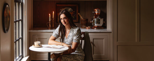 A woman sitting at a kitchen table near a window, enjoying a cup of coffee in a warm, inviting space with wooden cabinets and vintage decor.