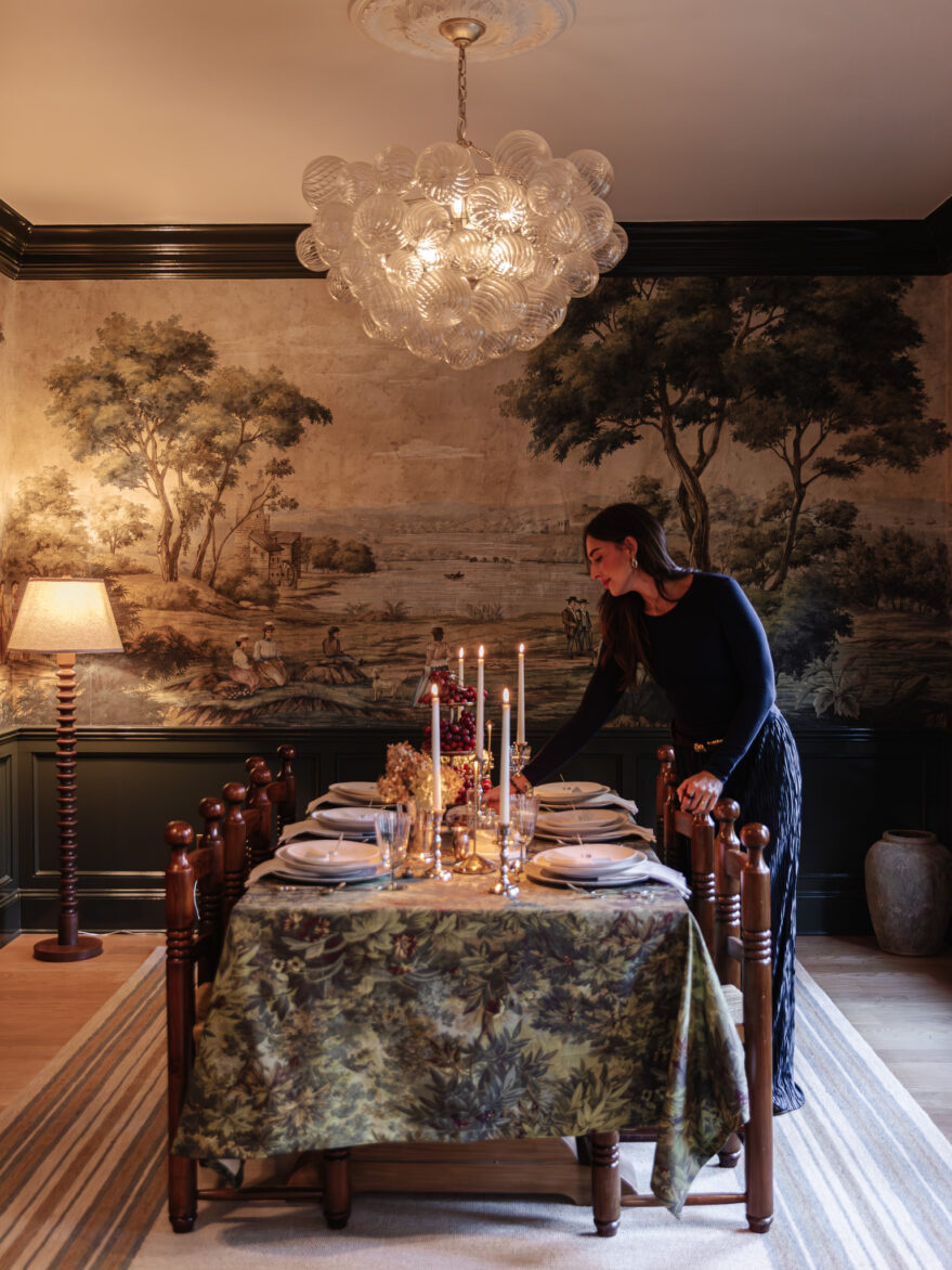 Elegant dining room with a mural wall, crystal chandelier, and a beautifully set table featuring candles and floral arrangements, showcasing classic interior design by Chris Loves Julia.