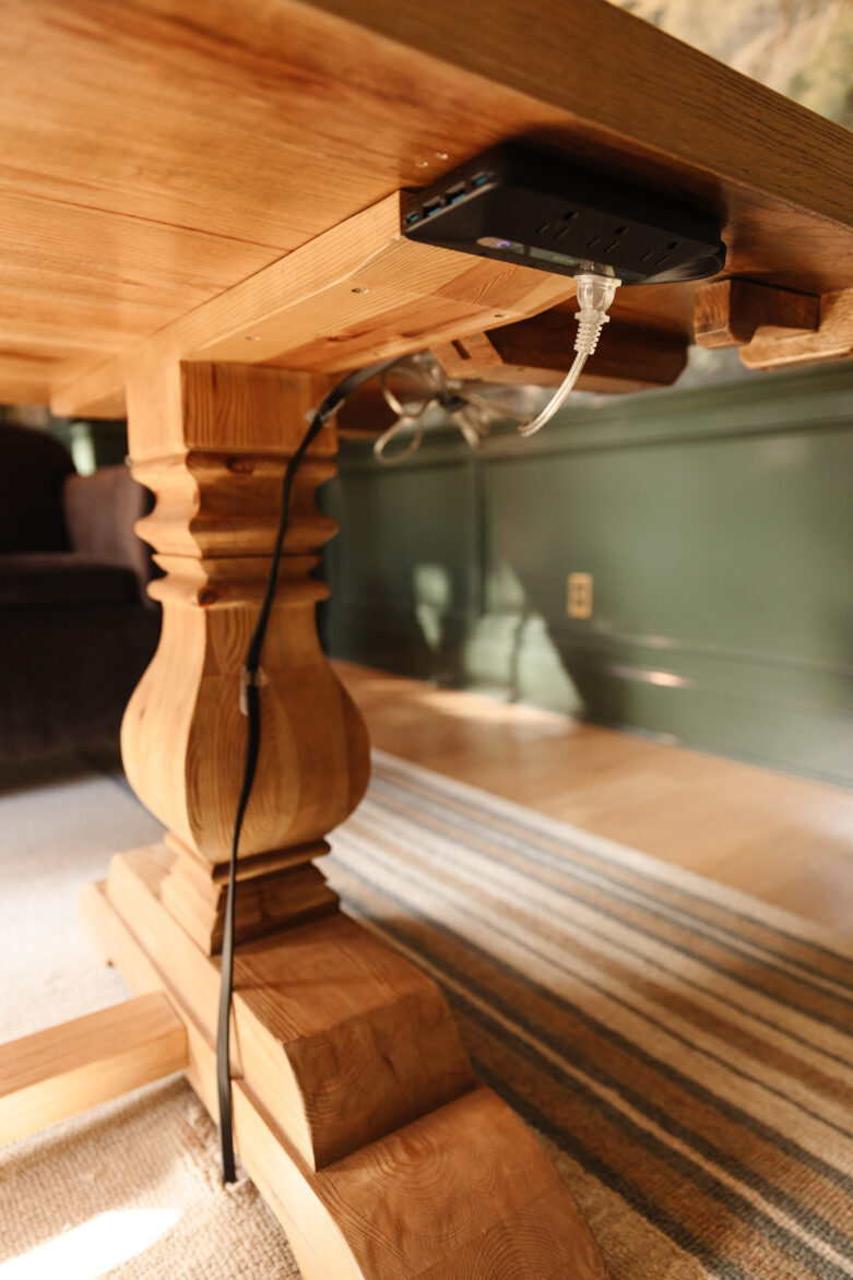 Close-up of wooden dining table turned desk with a power strip attached to the underside and a hole in the rug to feed the cable through