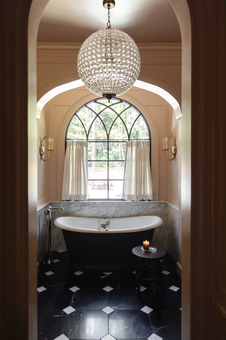 Luxurious bathroom featuring a classic freestanding bathtub beneath a large arched window with curtains, complemented by a stunning crystal chandelier and black-and-white checkered floor for a sophisticated look.
