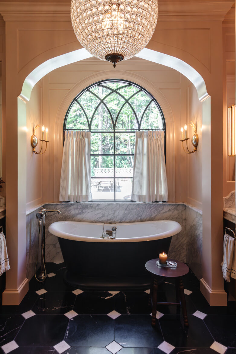 Luxurious bathroom with black and white marble accents, elegant bathtub beneath a stunning arched window, classic gold wall sconces, and a grand chandelier, showcasing home renovation by Chris Loves Julia.