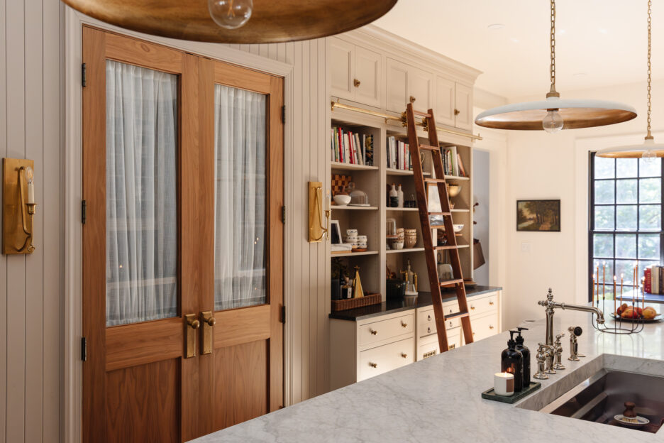 Warm wood-framed glass doors in a modern farmhouse kitchen featuring white beadboard cabinets, open shelving with cookbooks, a wooden ladder, and stylish pendant lighting, blending functionality with cozy aesthetic.