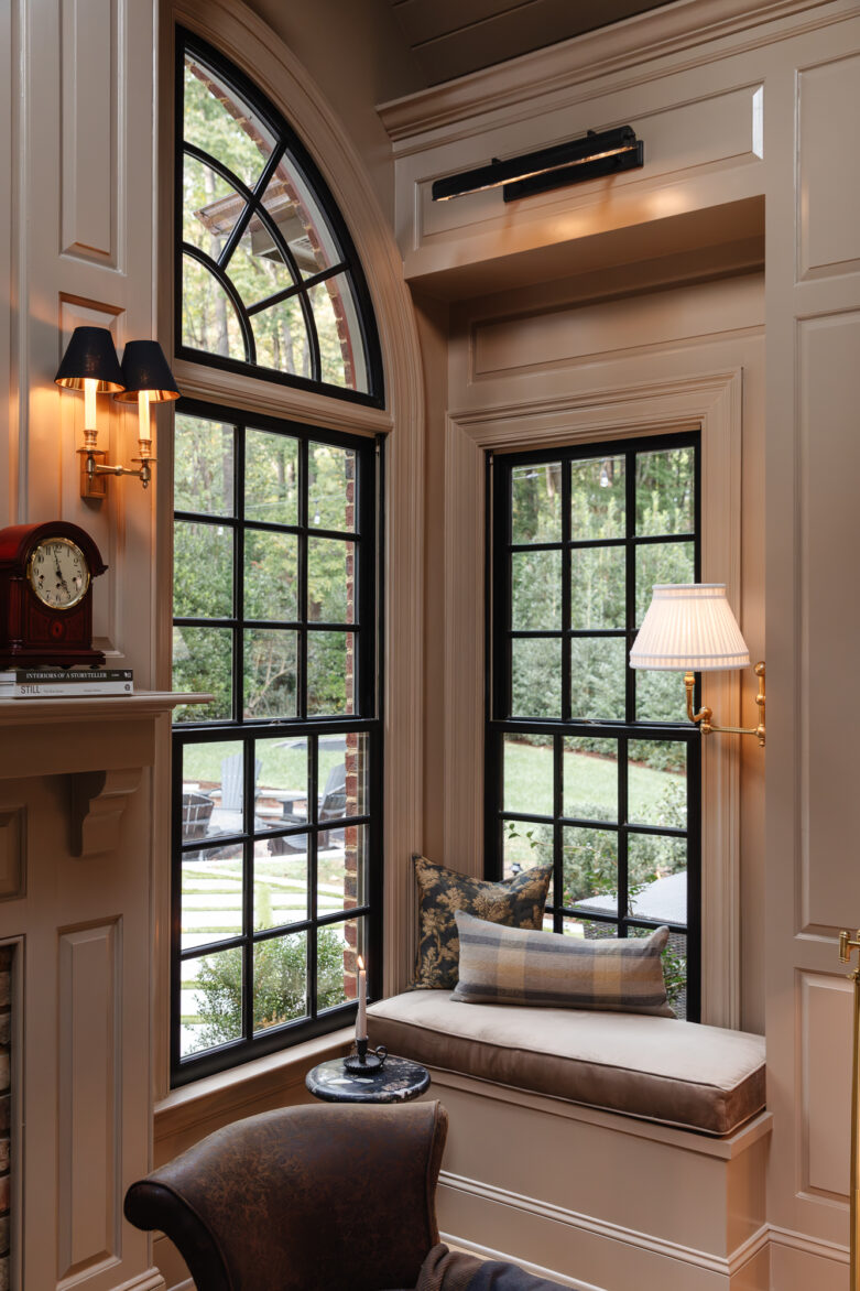 Elegant window seat nook with large black-framed windows, cozy cushions, and classic wall sconces, creating a warm and inviting reading corner in a sophisticated home interior.