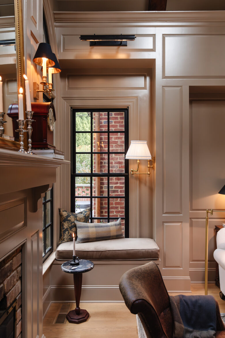 Cozy window seat with beige cushions, plaid pillows, and elegant wall sconces in a traditionally styled living room featuring cream-paneled walls and black-framed window, creating a warm and inviting space.
