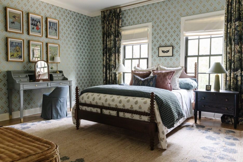 Cozy vintage-style bedroom with floral patterned wallpaper, wooden bed frame, and antique-inspired decor, featuring a vanity table, framed artwork, and natural light from large windows.