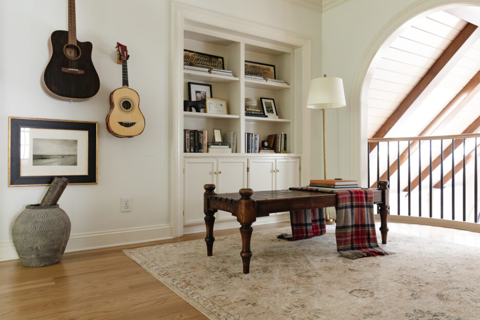 Cozy home library with a wooden coffee table, built-in bookshelf, decorative framed artwork, musical guitars on the wall, and a large arched window with wood beams, creating a warm and inviting atmosphere for reading and relaxing.
