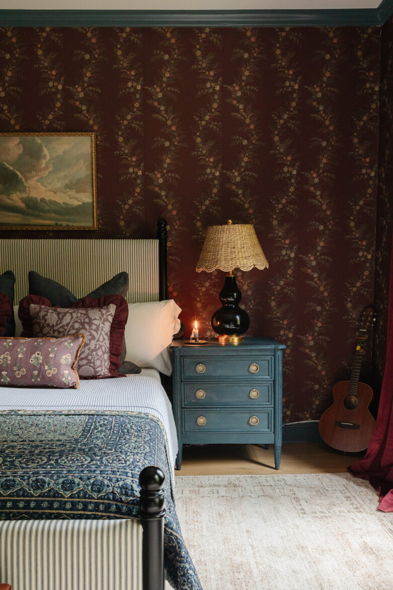 Versatile bedroom decor with vintage wallpaper and cozy bedding, accented by a teal nightstand, black lamp, and acoustic guitar, combining comfort and rustic charm for a stylish home interior.