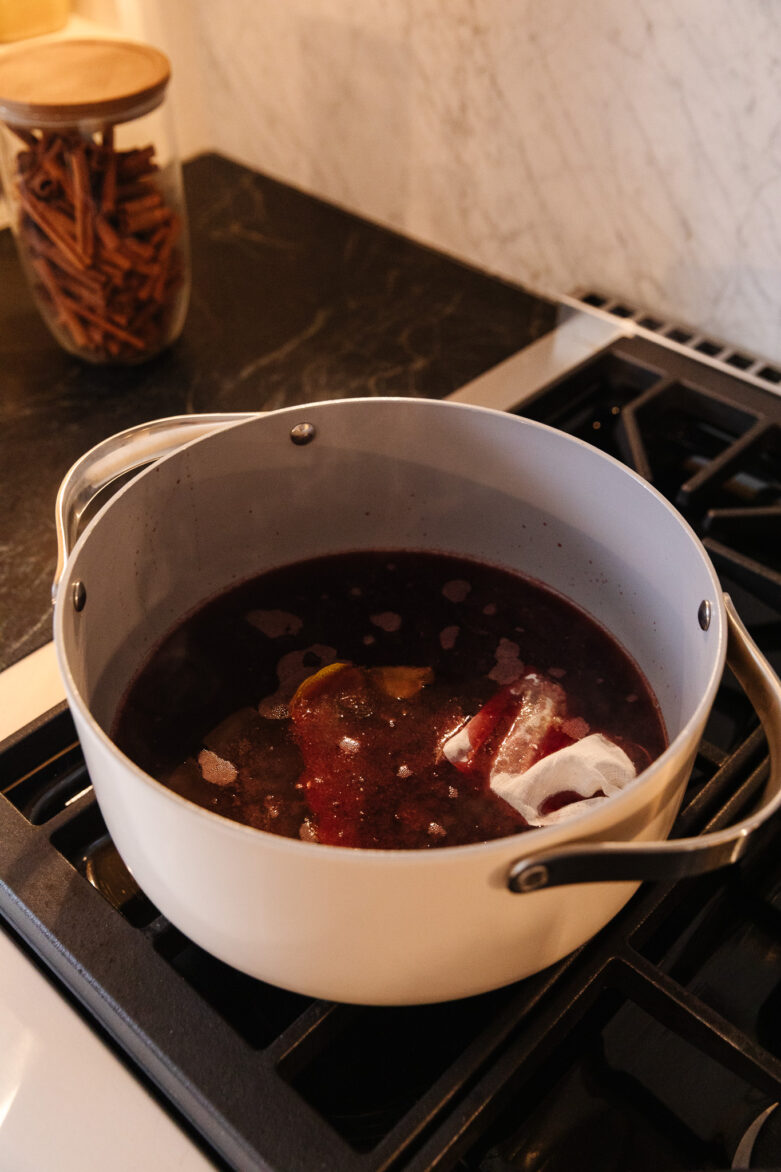 Brightly colored cranberry sauce cooking in a white pot on a stovetop, with ingredients like lemon peel and cinnamon sticks visible, ready to add festive flavor to holiday dining.