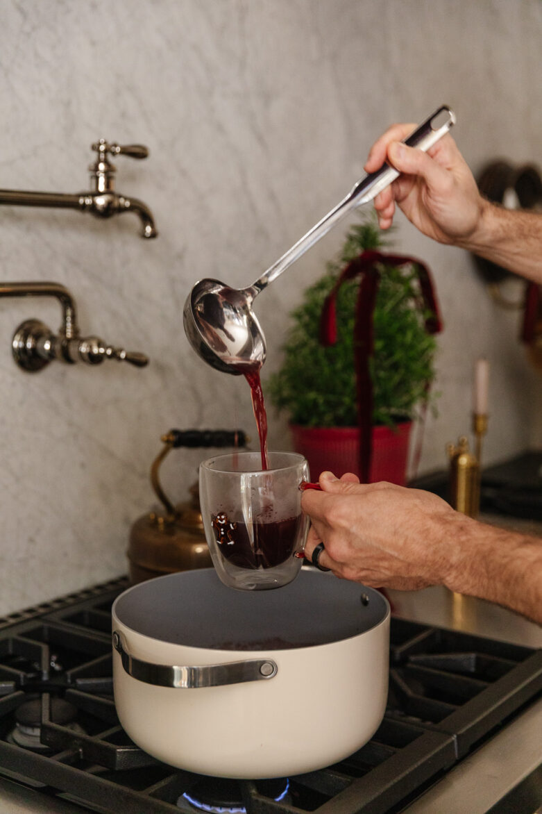 A person pouring red Glogg spiced drink from a ladle into a glass mug over a small stovetop, capturing a warm and inviting kitchen scene